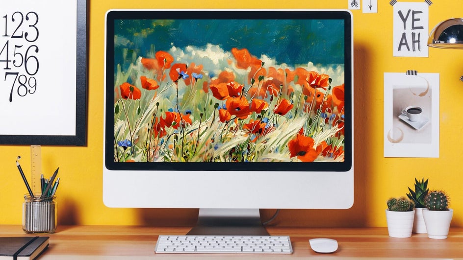 A computer monitor displays a vivid oil painting of red-orange poppies in a field. The desk below holds a plant, books, and more.